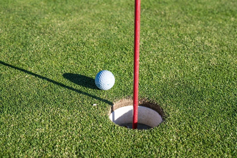 A red flagstick standing in the cup on a putting green, a golf ball at rest on the grass beside it in low raking sun.